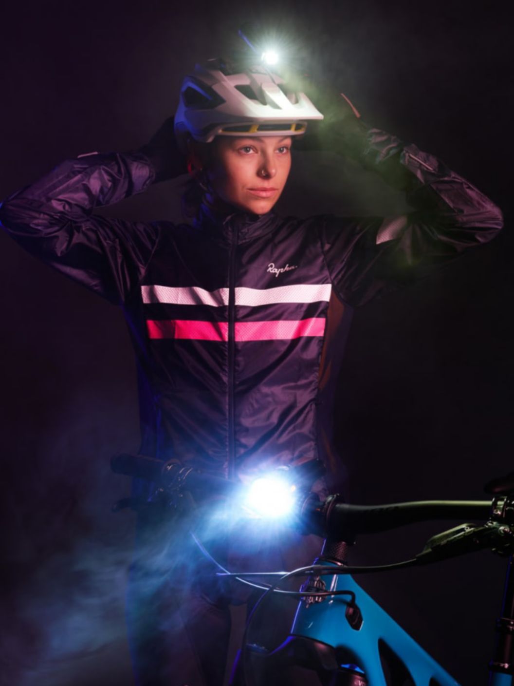 A rider in the dark wearing reflective riding apparel adjusts their helmet light while their handlebar mounted front light shines on light mist. 